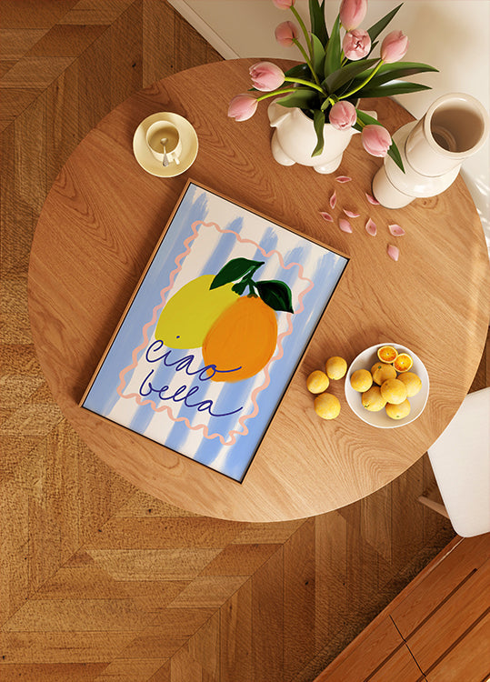 Ein Holztisch mit einem gerahmten Kunstwerk, das zwei Zitronen, eine Obstschale und eine Vase mit rosa Blumen zeigt.