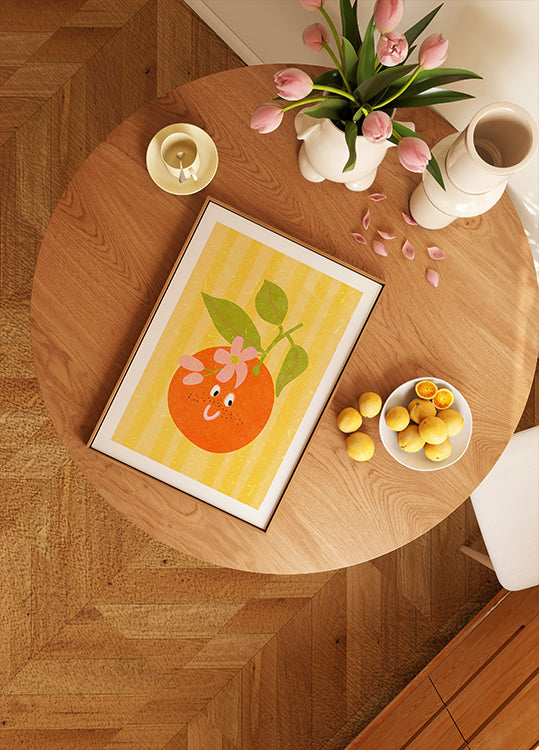 Ein Holztisch mit einem gerahmten Bild einer Orange, einer Schale mit Zitronen, einer Vase mit rosa Tulpen und einer Kerze darauf.